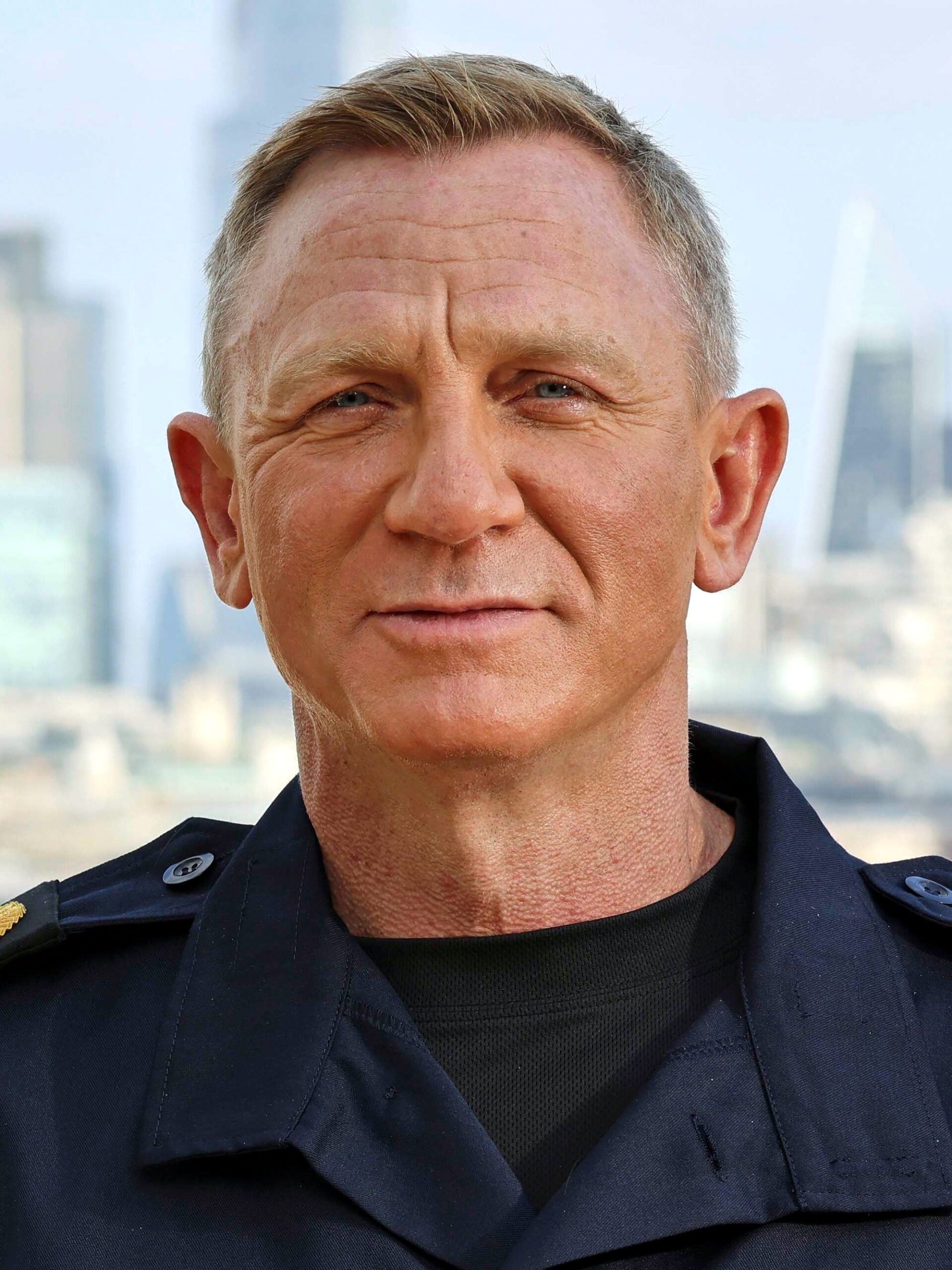 A man with short blond hair wearing a dark jacket stands outdoors with a city skyline blurred in the background.