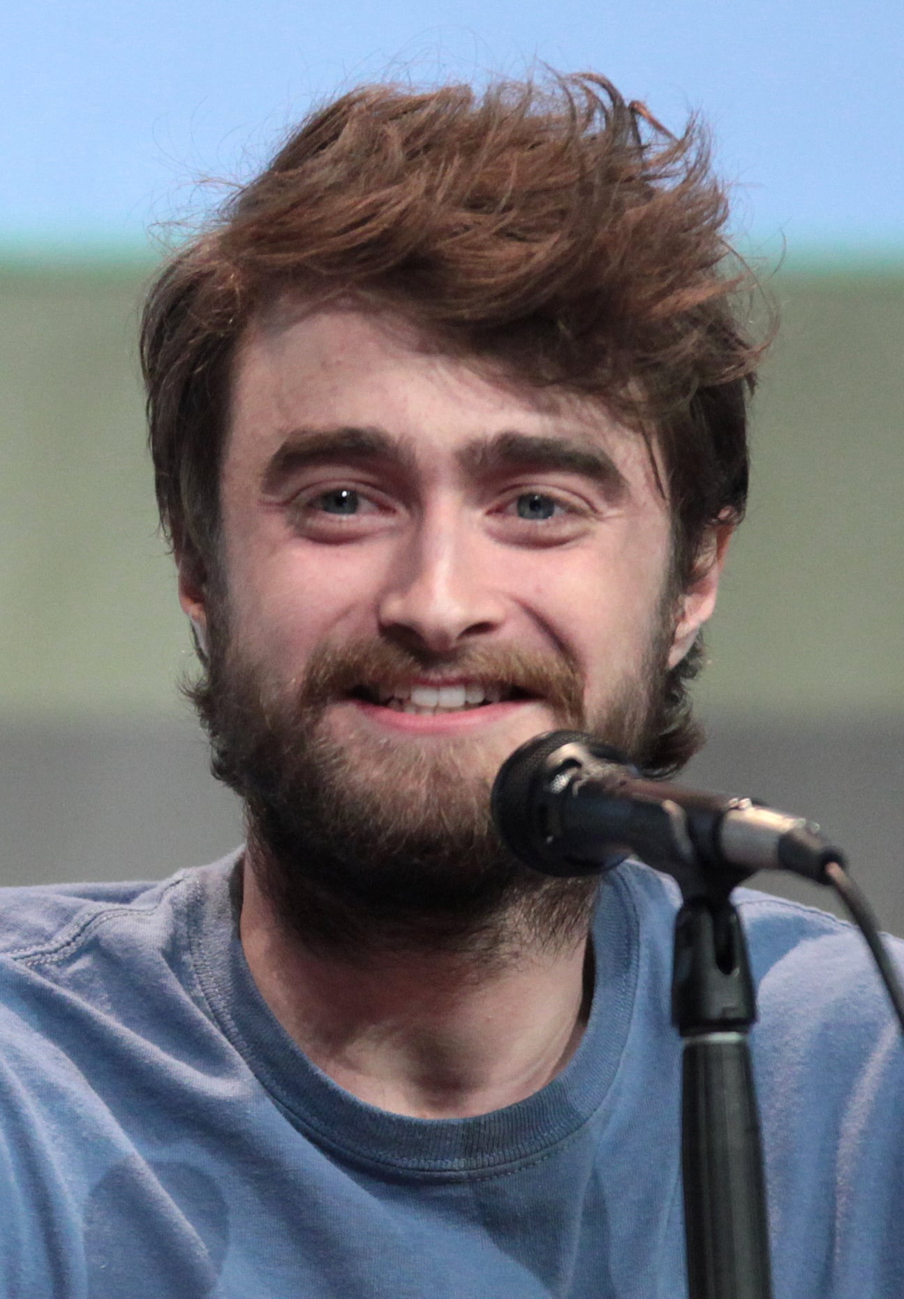 A man with tousled brown hair and a beard, wearing a blue shirt, smiles while speaking into a microphone at an event.