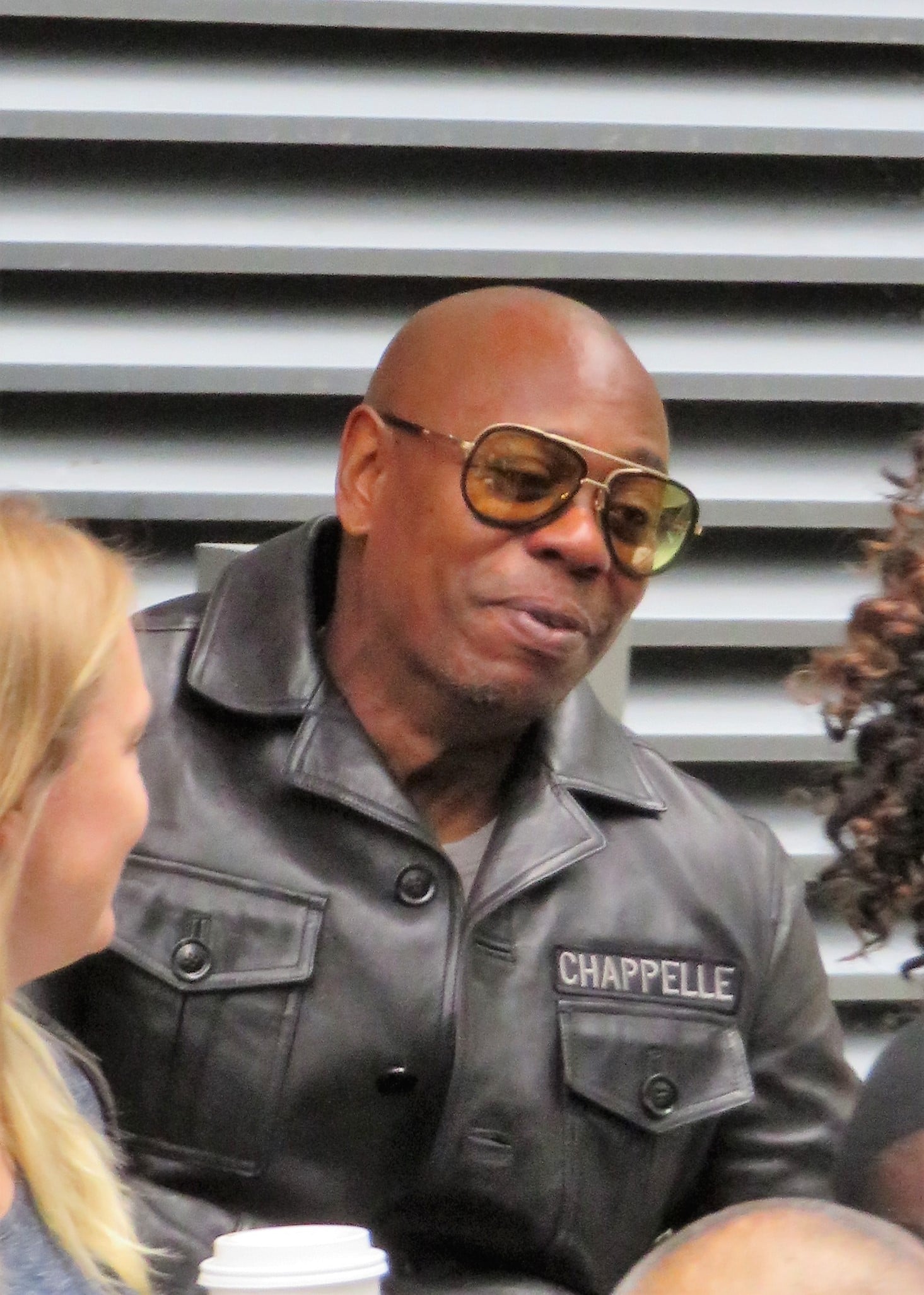 A man wearing a black leather jacket labeled "CHAPPELLE" and large sunglasses sits and talks with others near him, with a white coffee cup visible in the foreground.