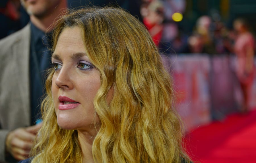 A woman with long, wavy blonde hair stands on a red carpet, looking to the side. She is wearing makeup and a dark outfit. The background is blurred with people and event banners visible.