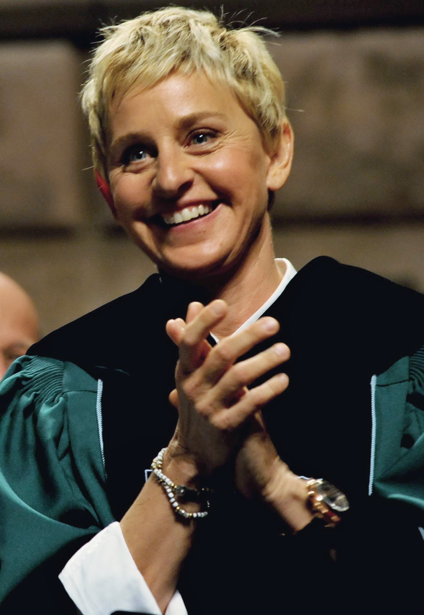 A person with short blonde hair, wearing a dark graduation gown with green trim, smiles and claps their hands during a ceremony.