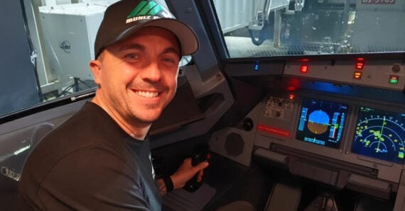 A smiling man wearing a black cap and t-shirt sits in an airplane cockpit, holding the control stick with flight instruments and screens visible in front of him.