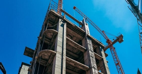 A multi-story concrete building under construction with red cranes towering above it against a clear blue sky.