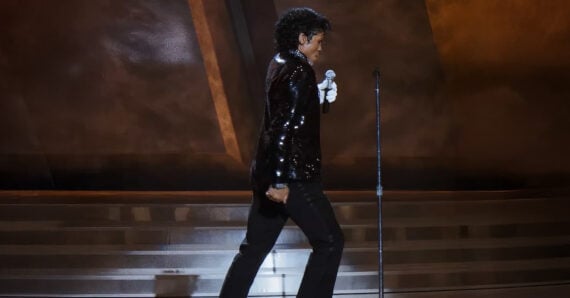 A performer in a black sequin jacket and black pants walks confidently across a stage, holding a white-gloved hand near a microphone stand, with dramatic lighting in the background.
