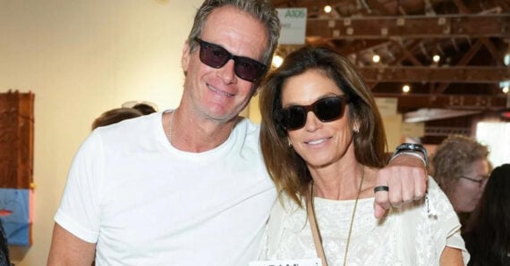 A man and woman wearing sunglasses and casual white clothing smile and pose together indoors at an event, with people and exposed wooden beams visible in the background.