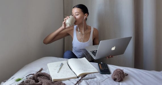 A person sits on a bed with a laptop, drinking from a glass. Open notebooks, a calculator, earphones, and yarn are on the bed, suggesting studying or working in a relaxed home setting.