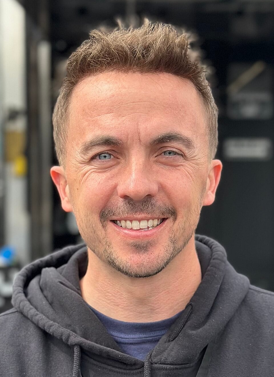 A man with short brown hair and blue eyes smiles at the camera, wearing a black hoodie. The background is blurred and dark, suggesting an outdoor or urban setting.