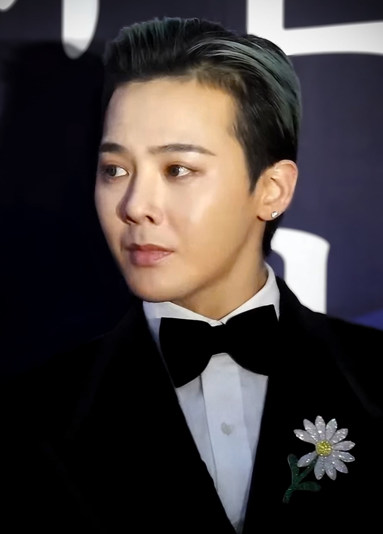 A person with slicked-back hair wearing a black tuxedo, bow tie, and a white shirt with a daisy flower brooch is posing against a dark background.