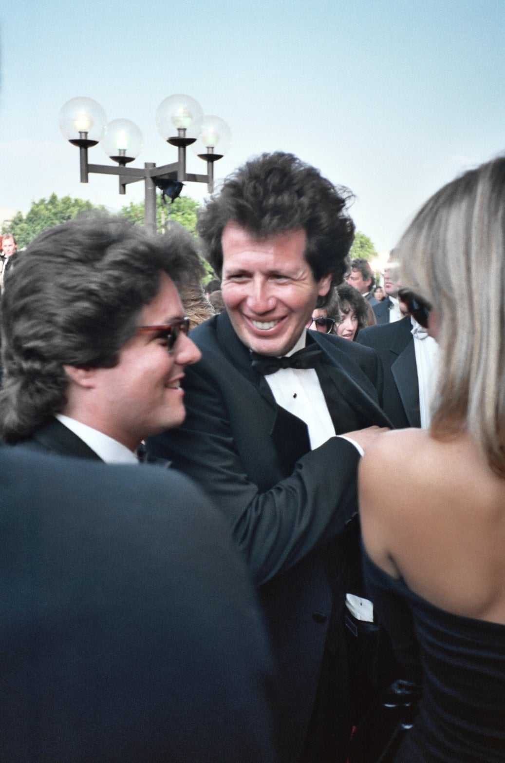 A man in a tuxedo smiles broadly while talking with others at an outdoor formal event. People around him are dressed in suits and dresses, and a lamp post is visible in the background.