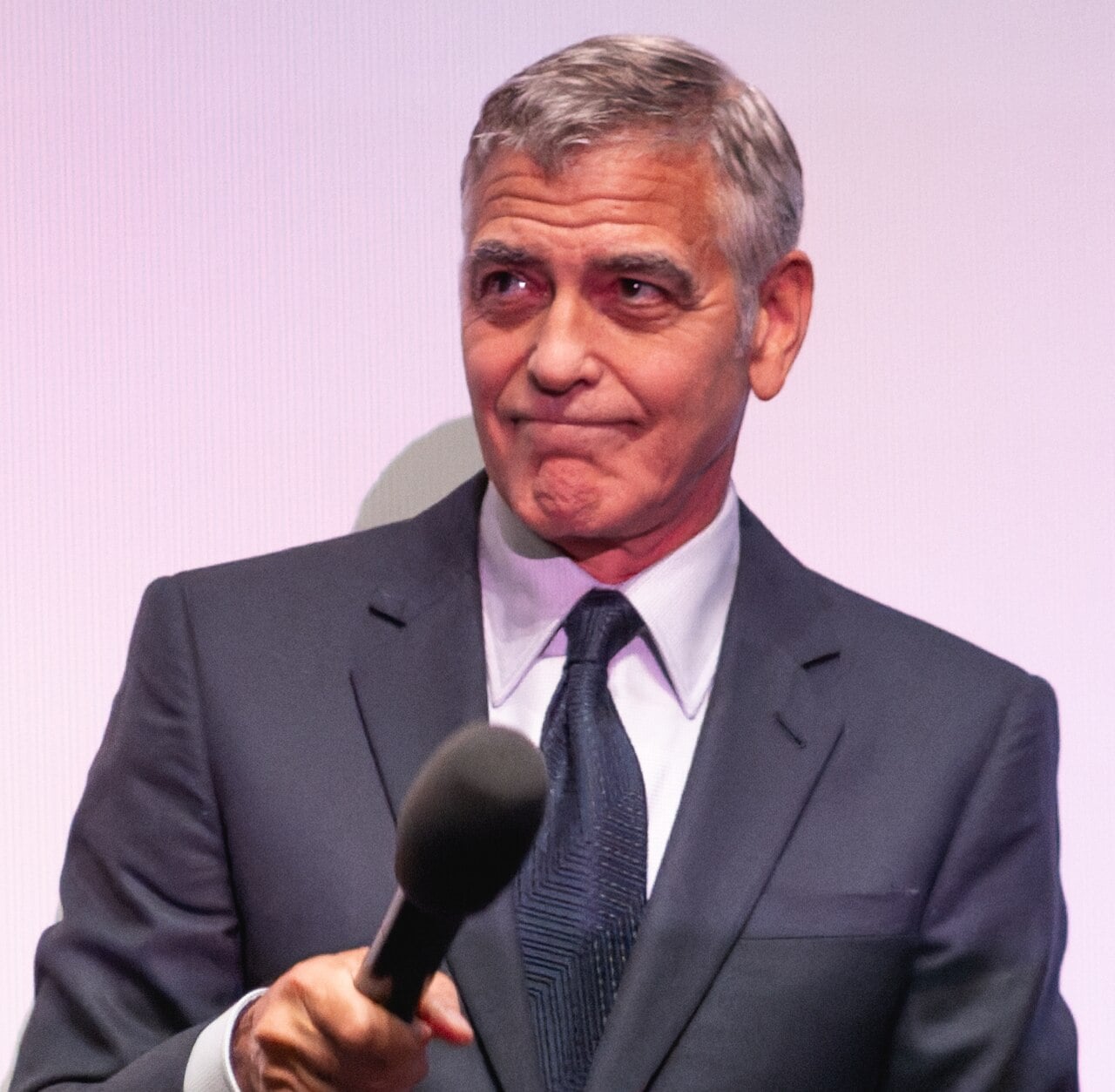 A man in a dark suit and tie holds a microphone, looking to the side with a slight smile against a light background.