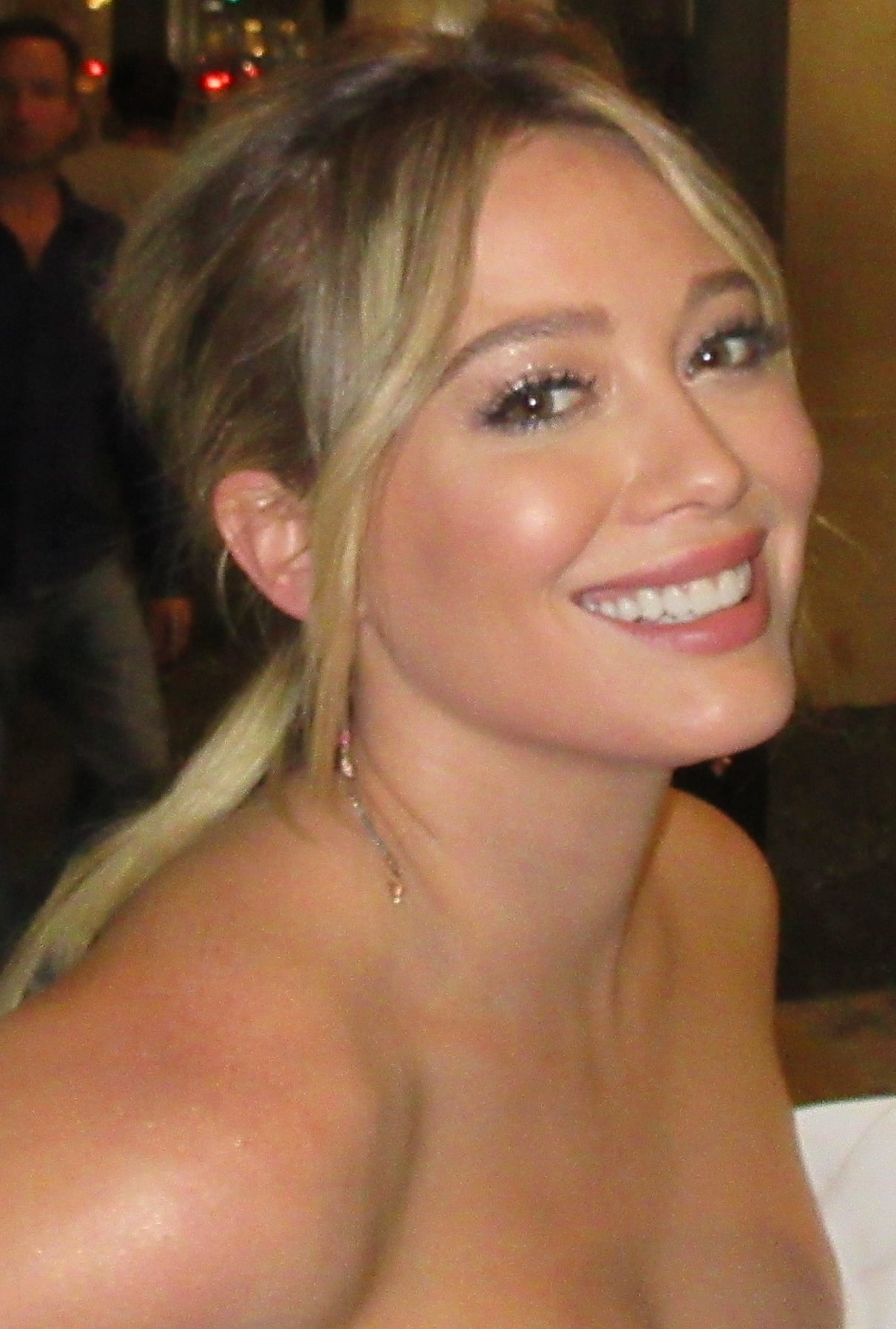 A woman with blonde hair styled in a loose ponytail smiles warmly at the camera. She is wearing subtle makeup and has bare shoulders, suggesting she may be dressed for a formal or special occasion.