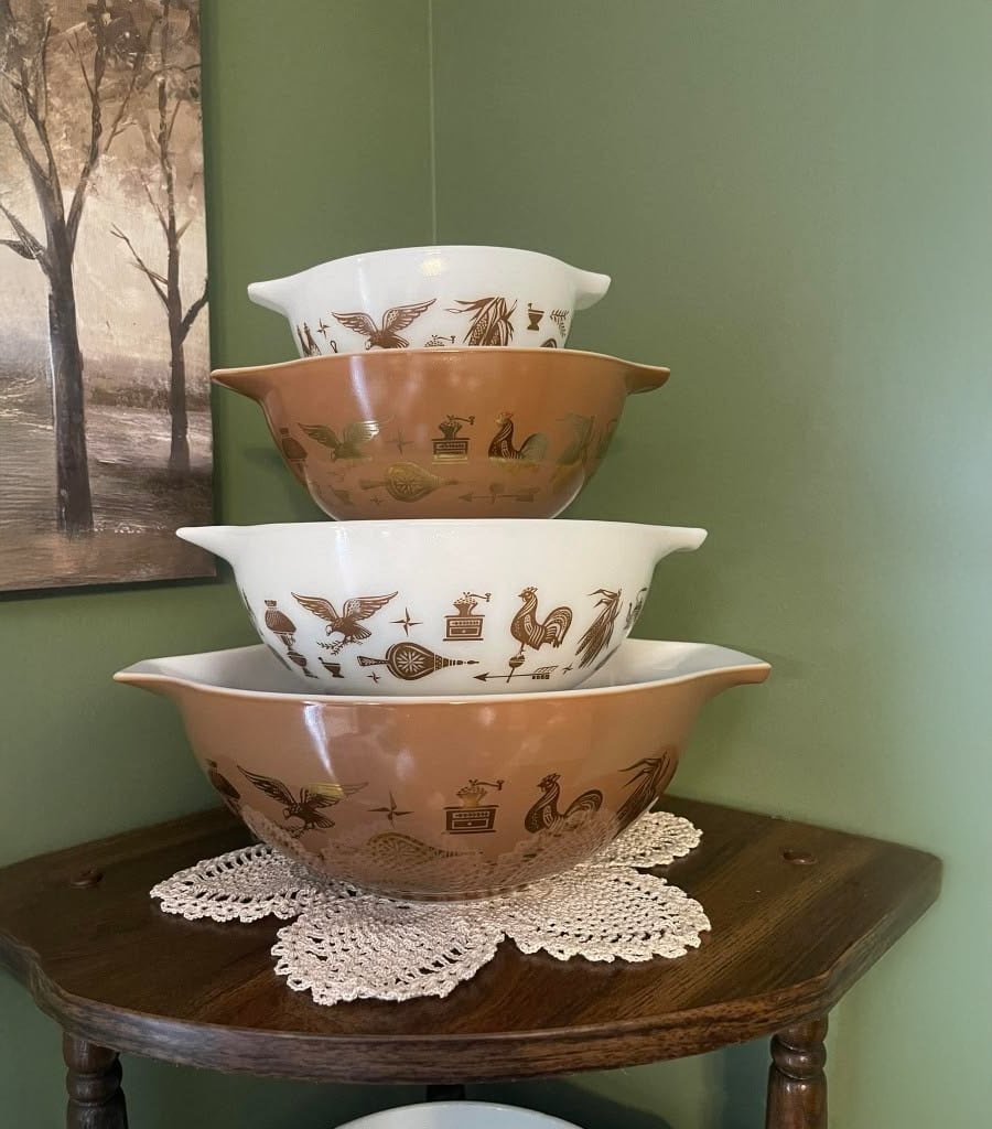 Three vintage mixing bowls with brown and white backgrounds and gold retro designs are stacked on a doily atop a wooden side table, against a green wall with a framed art piece on the left.