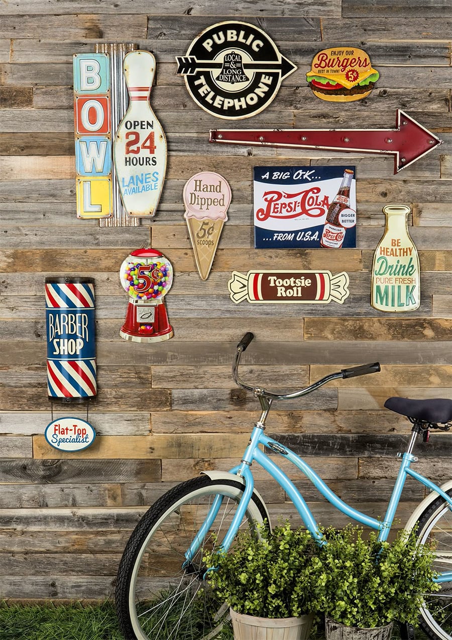 A rustic wooden wall decorated with colorful vintage-style metal signs, including a bowling alley sign, soda ads, a barber pole, a red arrow, and a Tootsie Roll. The front of a blue bicycle and a potted plant are in the foreground.