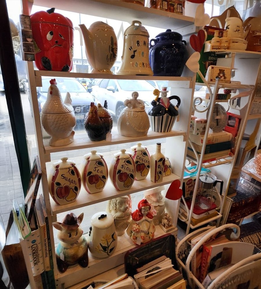 A display shelf in a store holding various ceramic teapots and jars shaped like animals and fruit, including chickens and a pig, labeled for tea, sugar, flour, and coffee, with other kitchenware and books nearby.