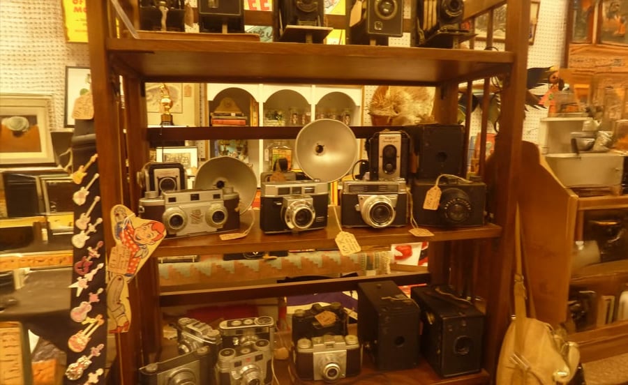 A wooden shelf displays various vintage cameras and camera equipment, including old box cameras and film cameras, with price tags attached. Other antique items are visible in the background.