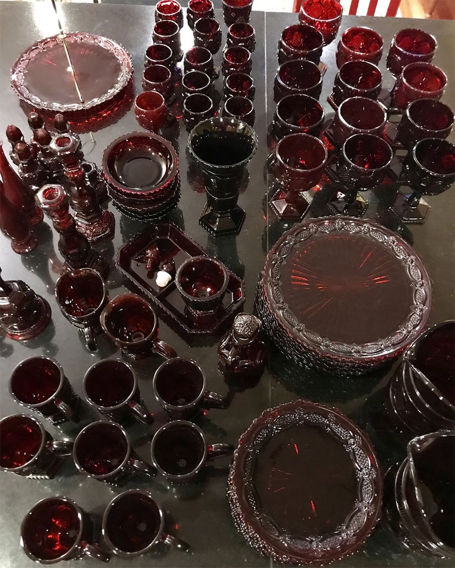 A collection of dark red vintage glassware, including plates, cups, goblets, bowls, and serving dishes, arranged neatly on a reflective black surface.