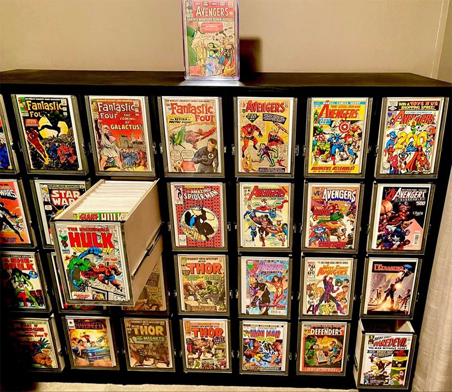 A black shelf displays a large collection of comic books, including titles like Fantastic Four, The Avengers, Thor, The Hulk, and The Defenders, with one drawer slightly open and a comic placed upright on top.
