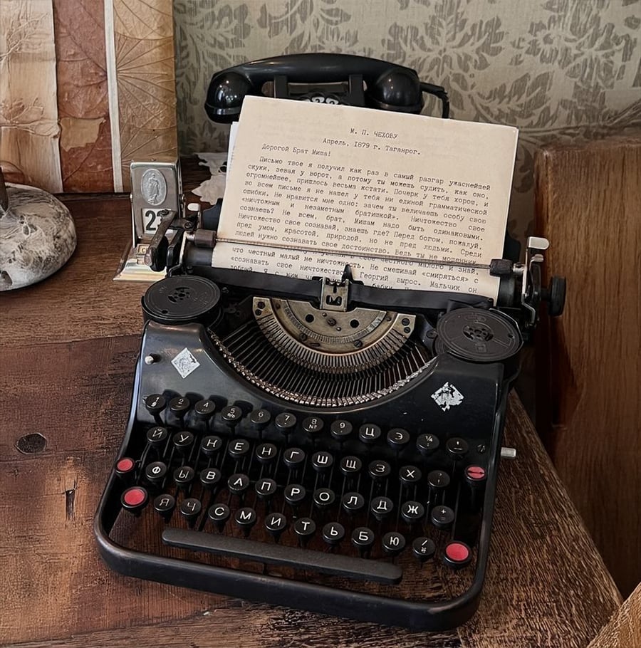 A vintage black typewriter with a Russian keyboard sits on a wooden desk. A sheet of typed Russian text is inserted, and an old rotary telephone rests behind the typewriter. Other antiques decorate the desk.