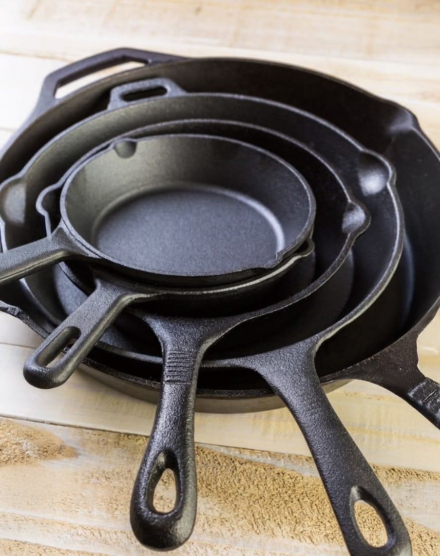 Four black cast iron skillets of different sizes stacked inside each other on a light wooden surface, with their handles extending outward.