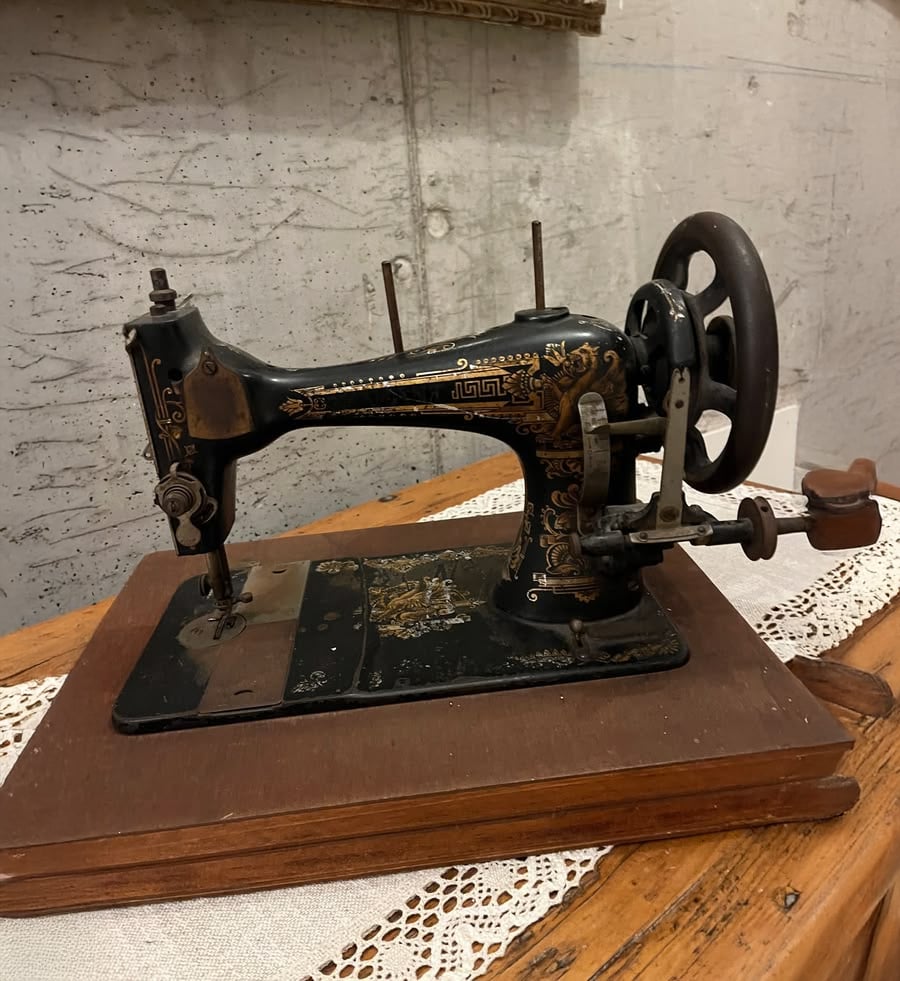 An antique black sewing machine with gold detailing sits on a wooden base atop a lace-covered table, against a textured gray wall.