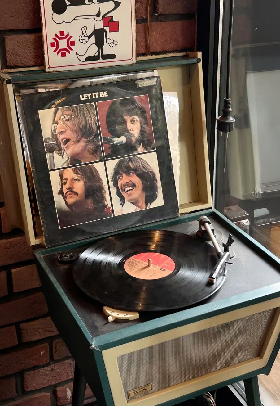 A vintage record player with The Beatles' "Let It Be" vinyl album cover propped up behind the spinning record. Four portraits of the band members are visible on the album cover.