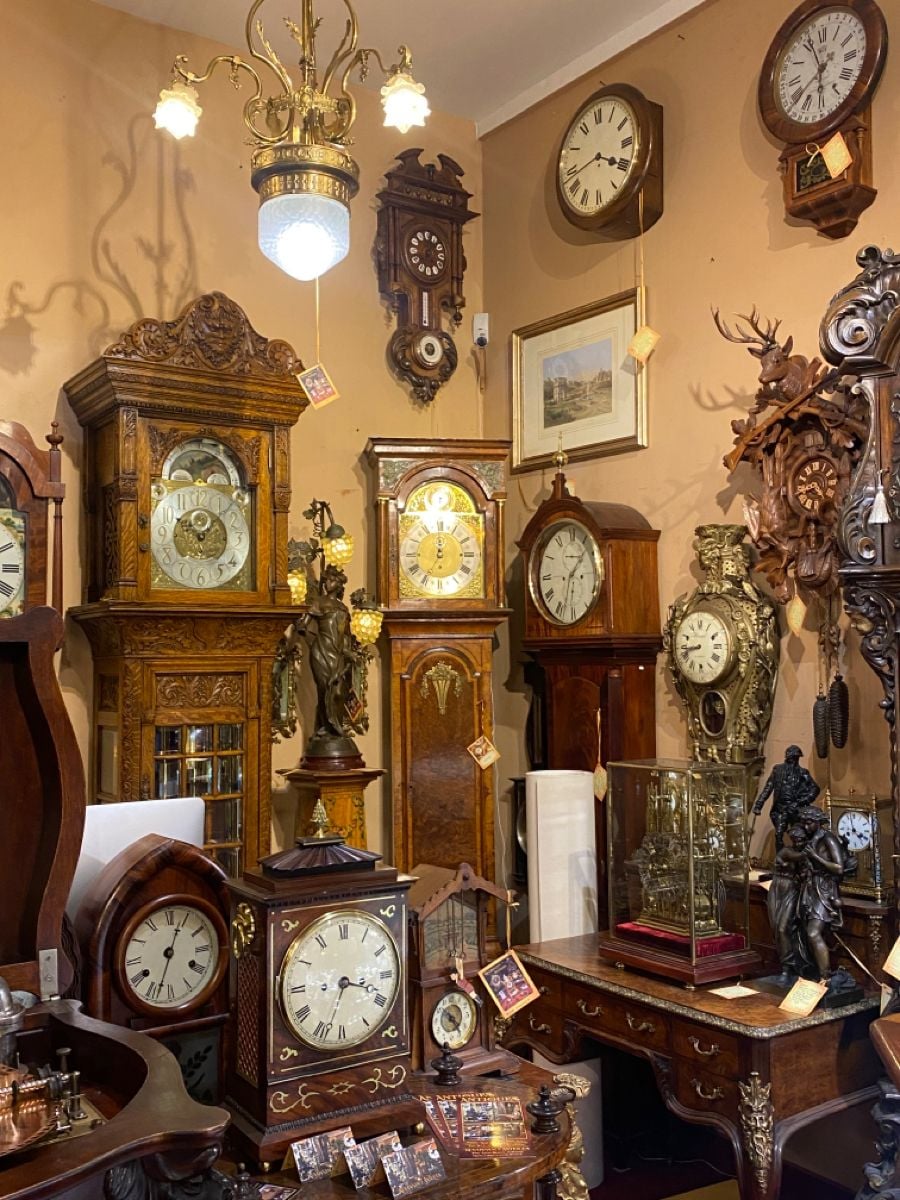 A room filled with various ornate antique clocks, including grandfather, mantel, and wall clocks, all displayed on and against wooden furniture. Warm lighting highlights the detailed craftsmanship and vintage decor.