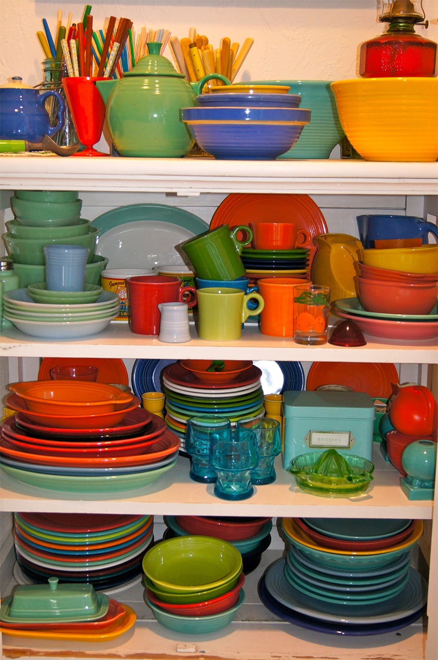 Colorful collection of vintage dishes, bowls, cups, plates, glasses, and pitchers arranged neatly on white shelves, featuring a vibrant mix of reds, blues, greens, oranges, yellows, and turquoise.