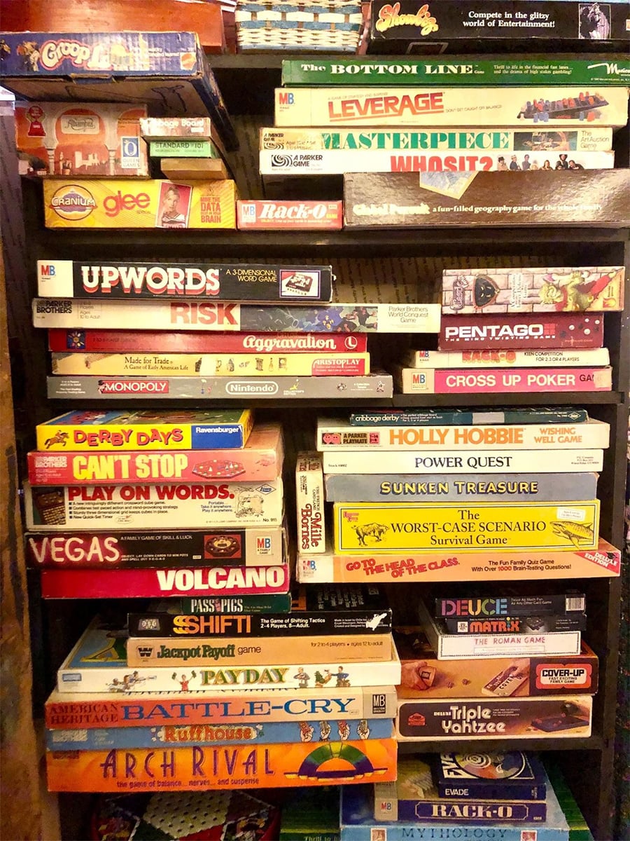 A cluttered shelf stacked with numerous board games, including titles like Risk, Monopoly, Masterpiece, Vegas, Upwords, Aggravation, and others, with colorful and varied game boxes tightly packed together.