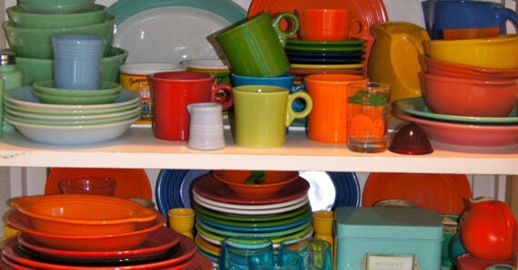 A shelf filled with brightly colored dishes, bowls, cups, and glasses in various shapes and sizes, stacked and arranged closely together. The tableware is mostly orange, green, yellow, red, and blue.