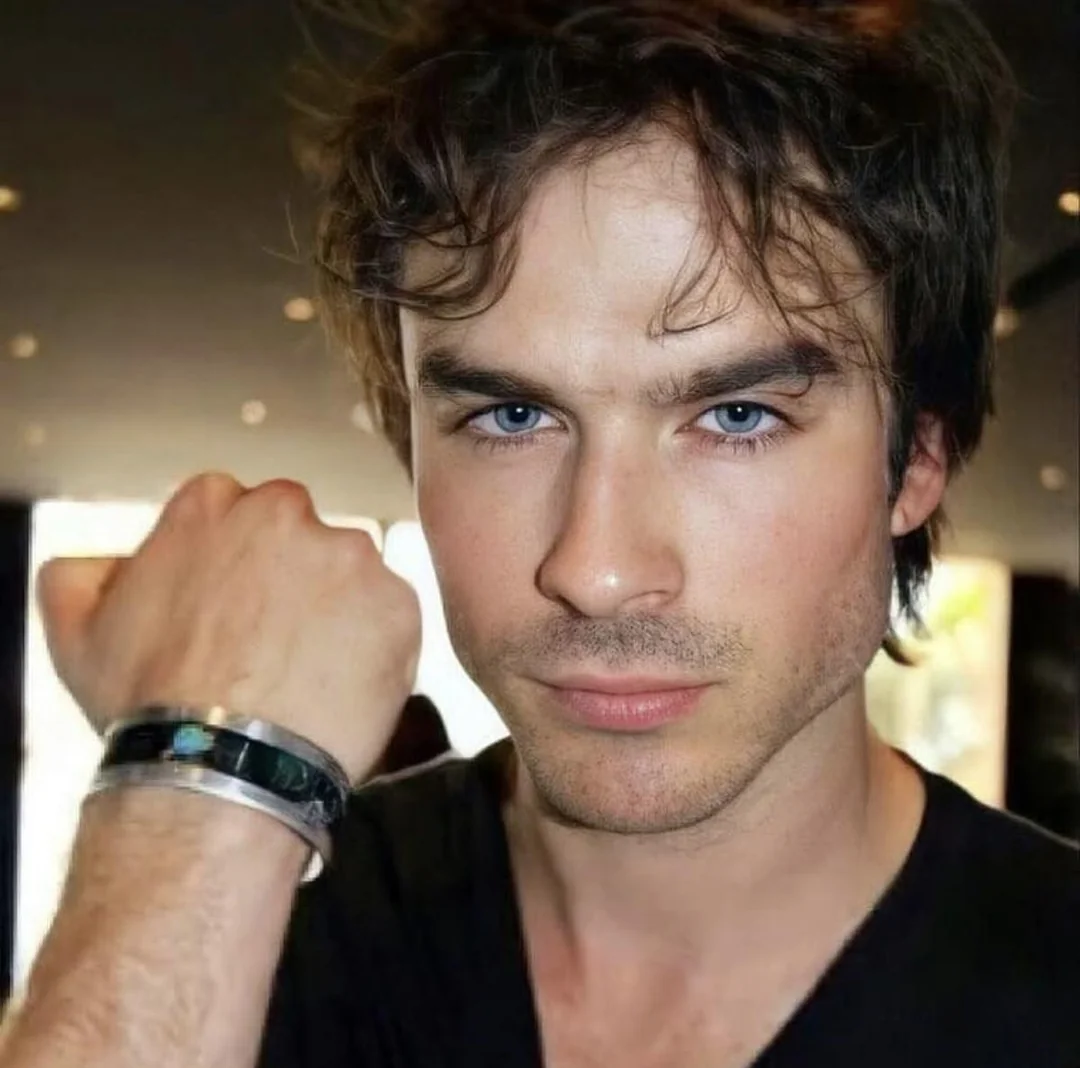 A man with tousled brown hair and blue eyes looks directly at the camera, holding up his fist to display a black and silver bracelet. He is wearing a black shirt in a softly lit indoor setting.