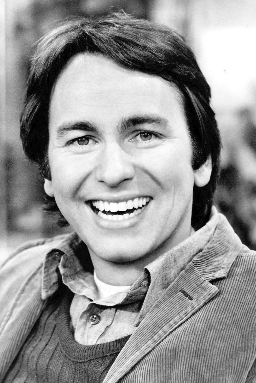 A man with medium-length dark hair is smiling widely. He is wearing a corduroy jacket over a sweater and collared shirt. The image is black and white, and the background is blurred.
