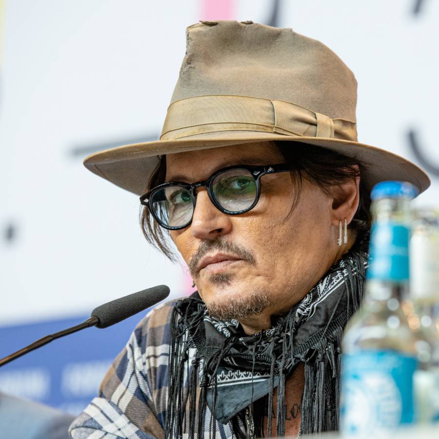 A man wearing a tan hat, dark glasses, plaid shirt, and a patterned scarf sits at a table with microphones and water bottles, speaking at a press event.