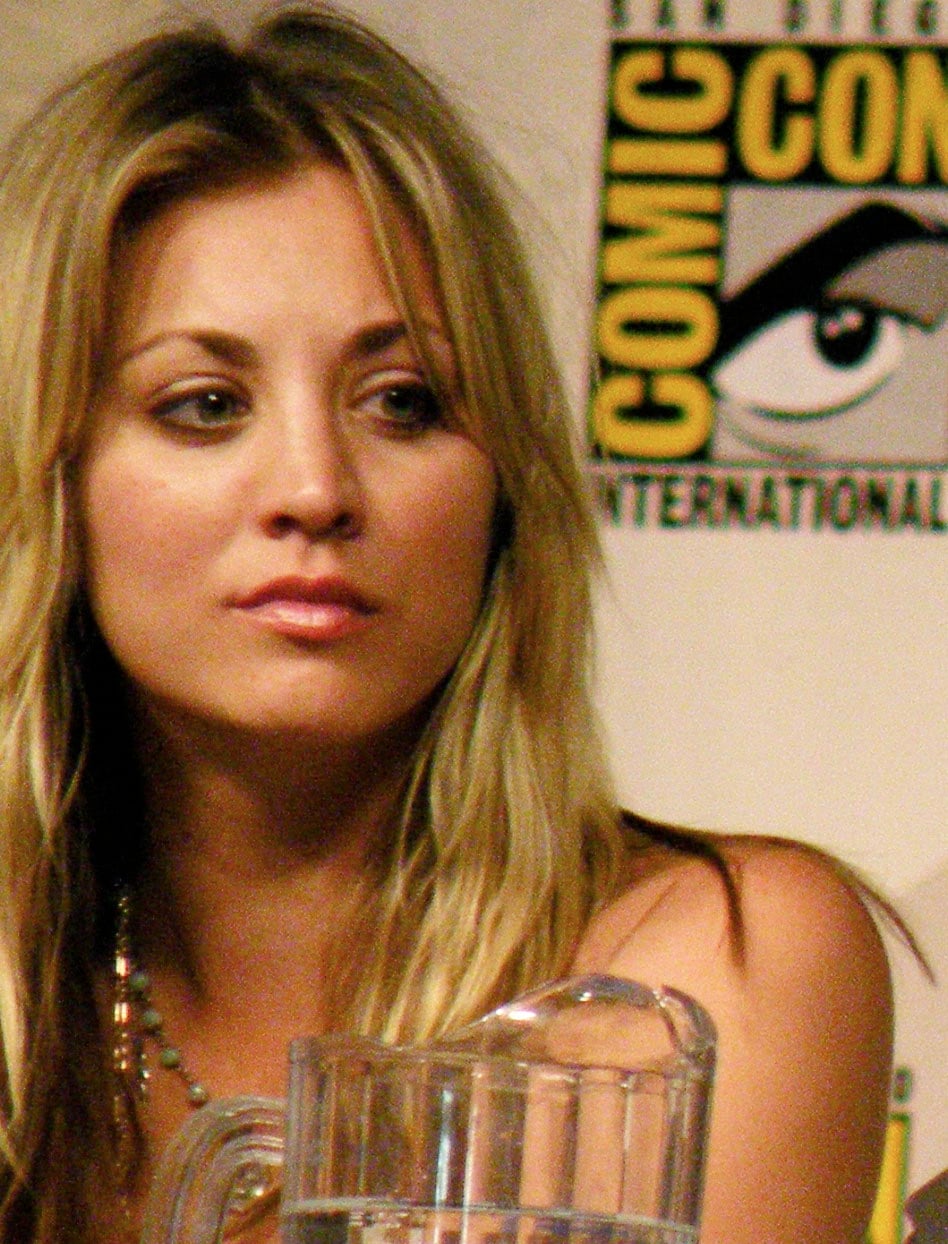 A woman with long blonde hair sits in front of a Comic-Con International banner, looking slightly to the side. A clear pitcher is visible in the foreground.