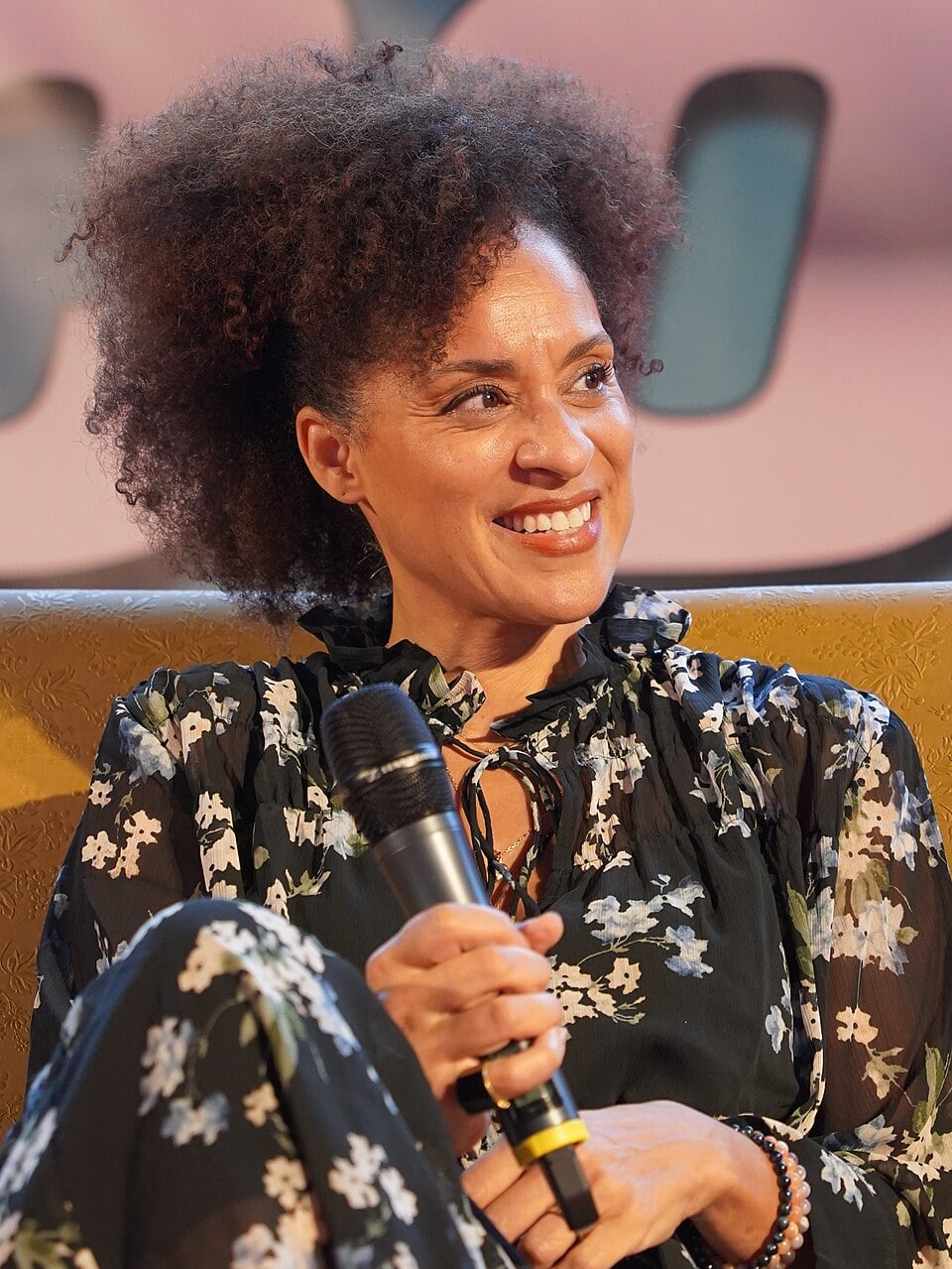 A smiling woman with curly hair sits on a yellow patterned couch, holding a microphone. She is wearing a dark floral dress and looking to the side. The background is blurry with large white shapes.