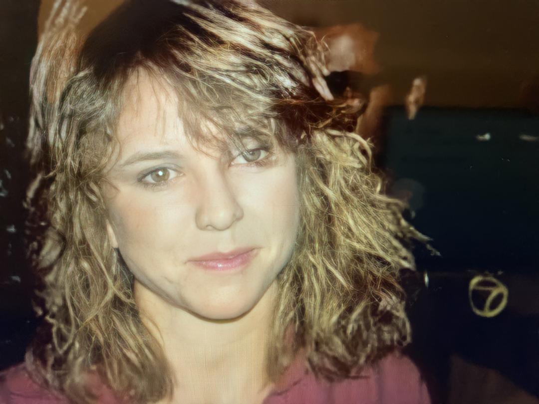 A woman with wavy, light brown hair and light eyes smiles softly at the camera, wearing a mauve top. The background is dark and blurry.