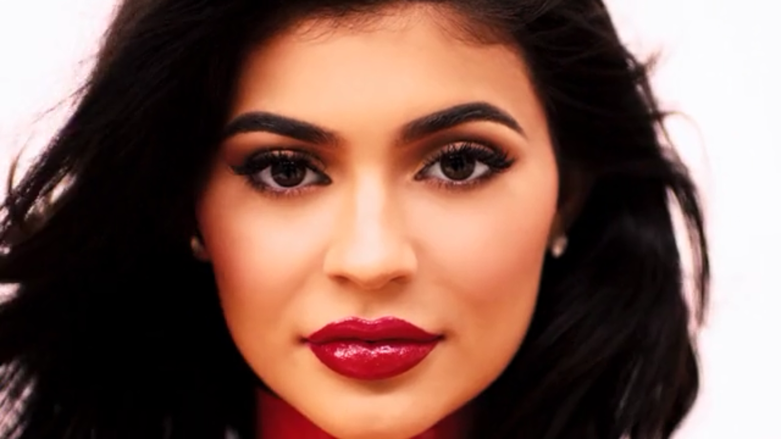 A woman with long dark hair, bold eyebrows, dramatic eye makeup, and glossy red lips looks directly at the camera against a light background.