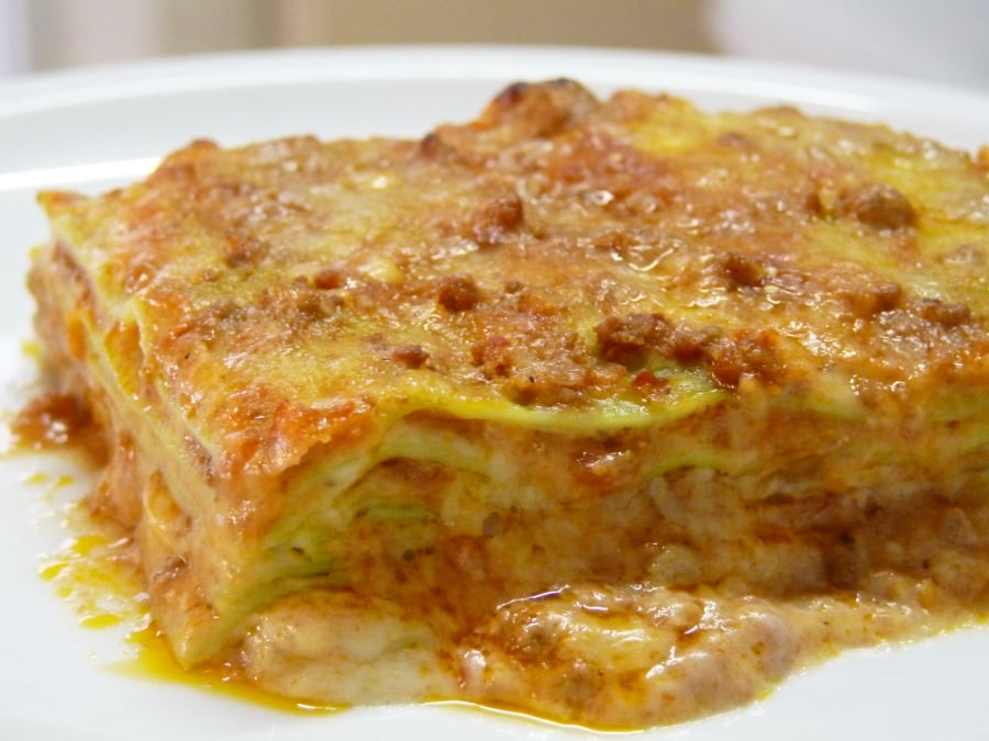 A close-up of a slice of lasagna on a white plate, showing layers of pasta, melted cheese, and a rich meat and tomato sauce.