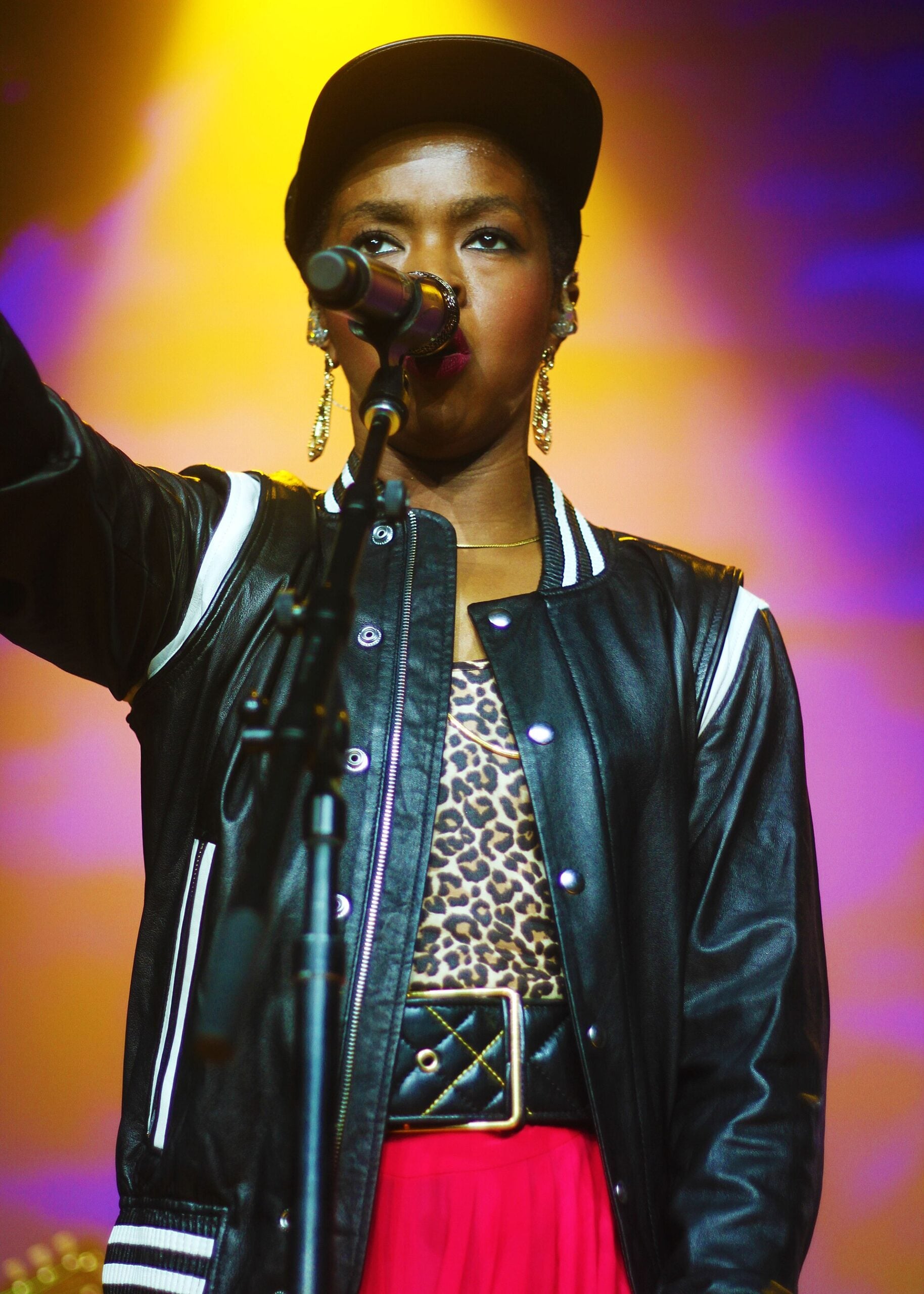 A woman sings into a microphone on stage, wearing a black cap, hoop earrings, a black varsity jacket, a leopard print top, and a red skirt, with dramatic yellow and purple stage lighting behind her.
