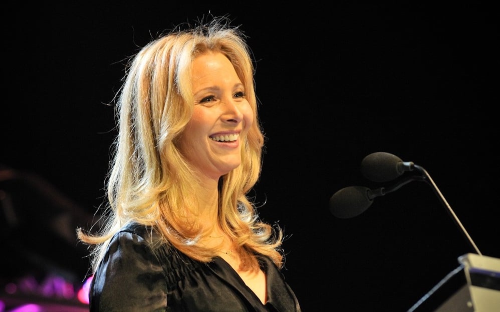A woman with long blonde hair smiles while standing at a podium and speaking into a microphone. She is wearing a black top and the background is dark.