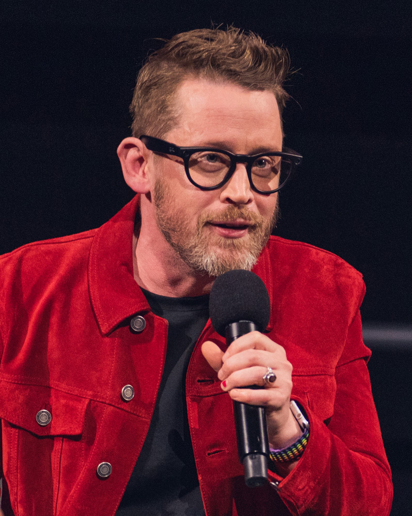 A man with short hair and a beard, wearing black glasses and a red jacket, holds a microphone while speaking.