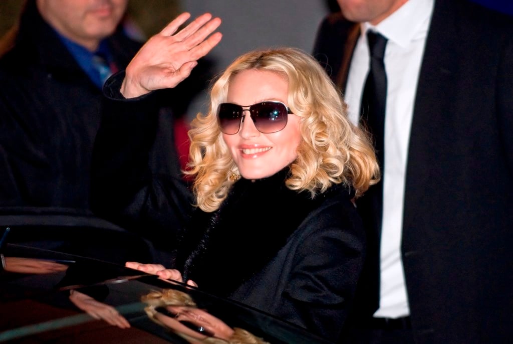 A woman with blonde curly hair and sunglasses smiles and waves while getting out of a car, surrounded by people in formal attire.