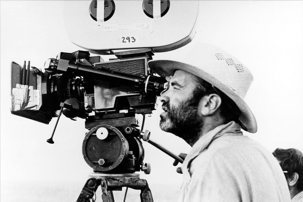 A man wearing a hat closely peers through a film camera’s viewfinder, focusing intently. The camera is large and mounted on a tripod. The background is plain and out of focus. The image is in black and white.
