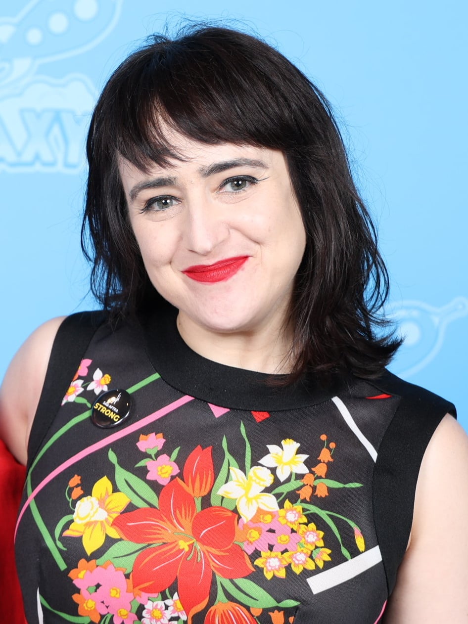 A woman with short dark hair and red lipstick smiles in front of a blue background. She wears a sleeveless black dress with a bright floral pattern and a pin that says "STRONG.