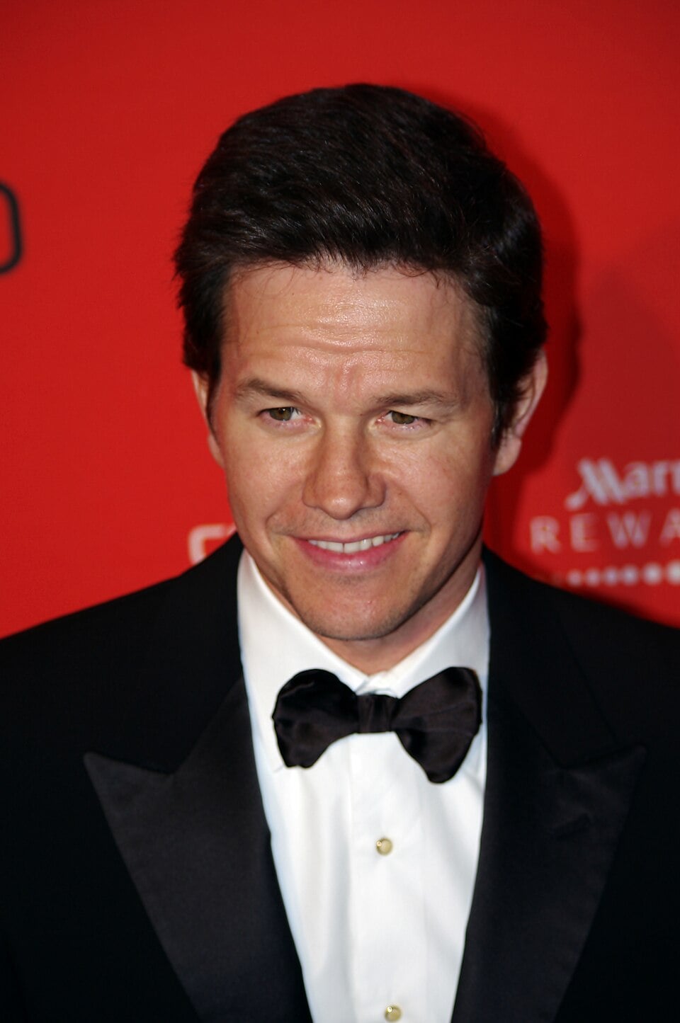 A man with short dark hair wearing a black tuxedo, white shirt, and black bow tie smiles in front of a red backdrop with white text and logos.