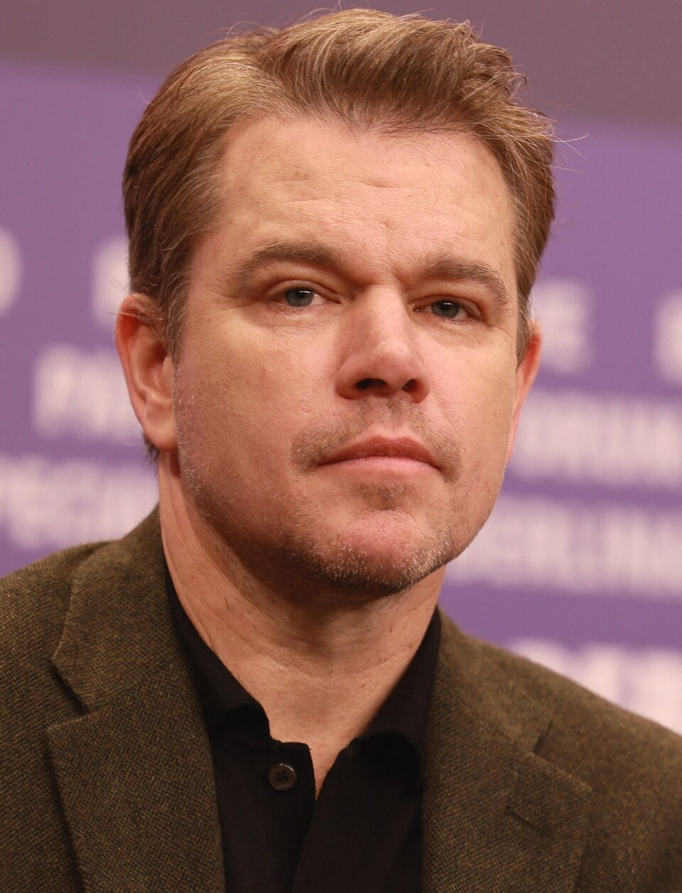 A man with short, light brown hair and light facial stubble wearing a dark jacket and black shirt, looking forward with a neutral expression in front of a blurred background.