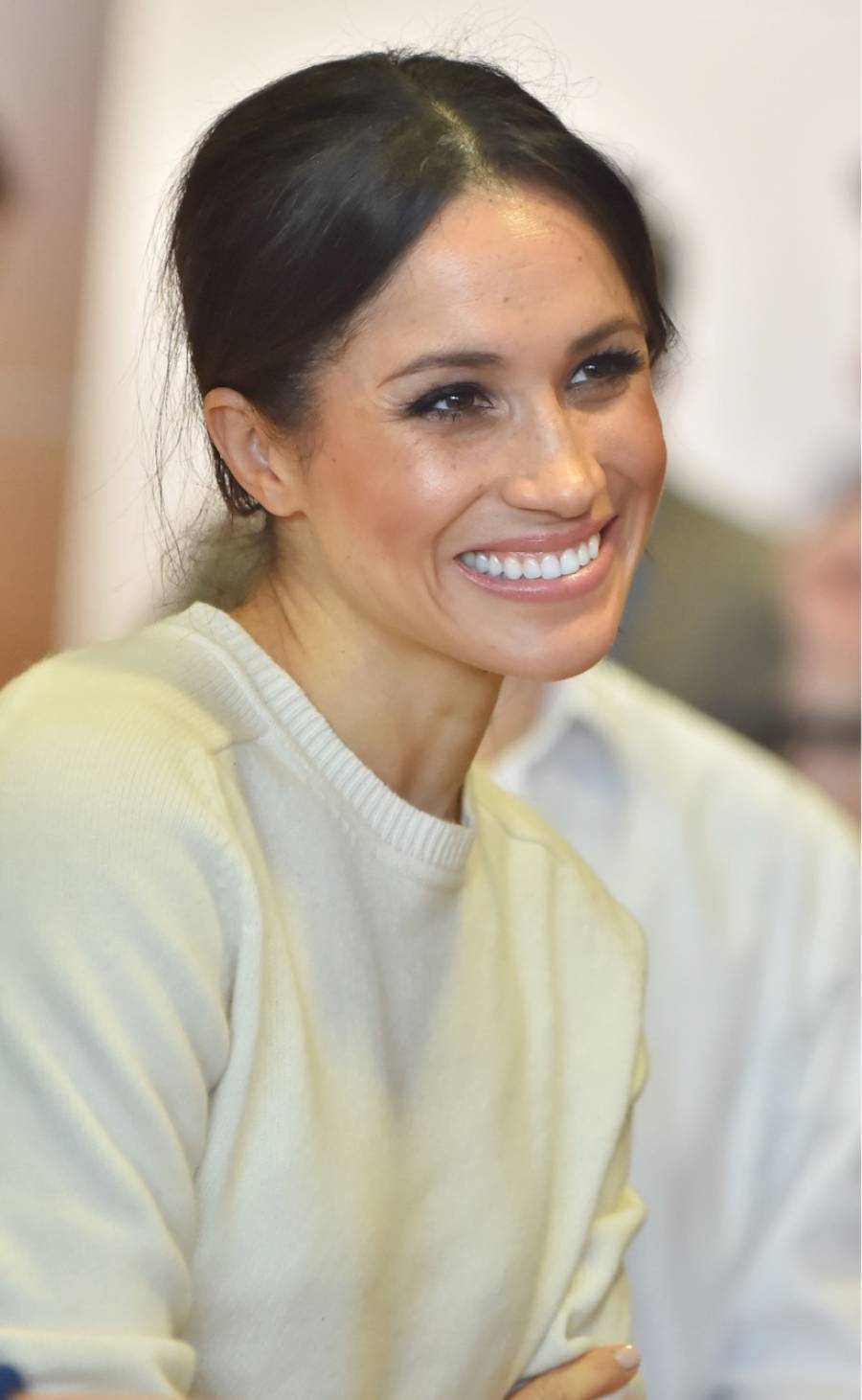 A woman with dark hair tied back, wearing a light-colored sweater, smiles warmly while looking slightly to the side. The background is softly blurred.