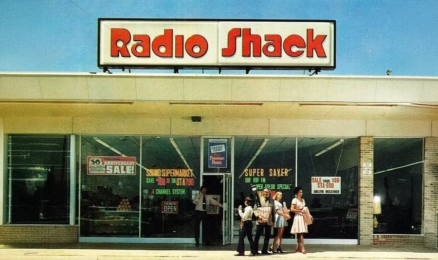 A group of five people stand outside a vintage Radio Shack store with large front windows displaying sale signs. The store's bold red sign is prominent above the entrance.