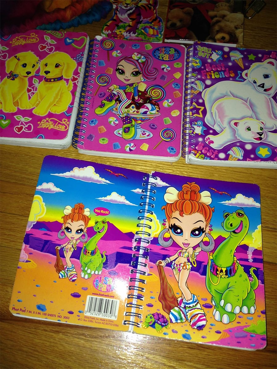 Four colorful spiral notebooks are displayed on a wooden surface, featuring cartoon designs: puppies, a girl with rainbow hair, polar bears, and a girl with a green dinosaur in a desert scene.