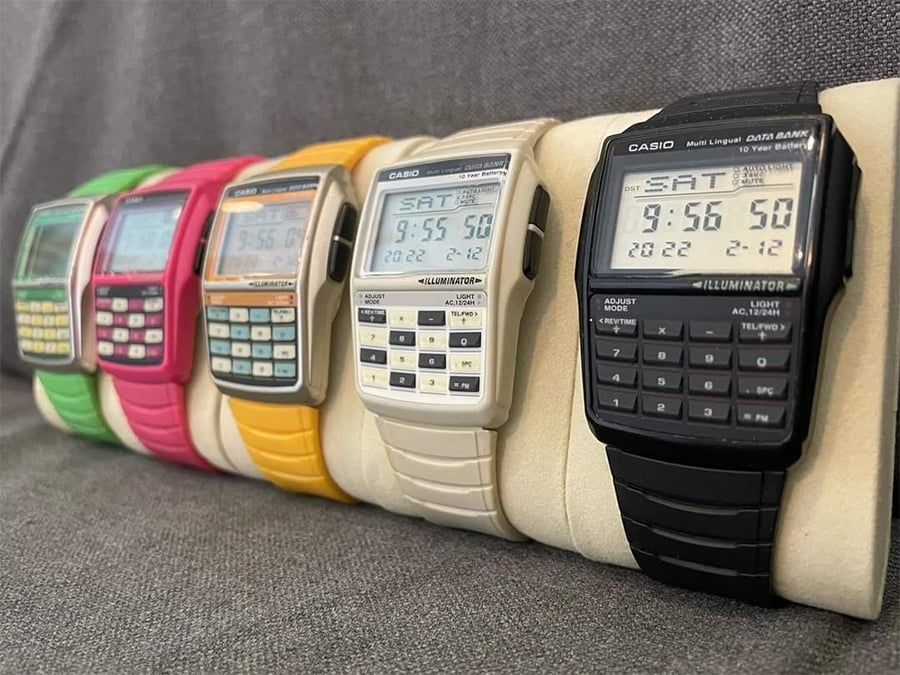 Five Casio calculator watches in green, pink, blue, yellow, white, and black bands are displayed on a gray background, all showing the same time and date on their digital screens.