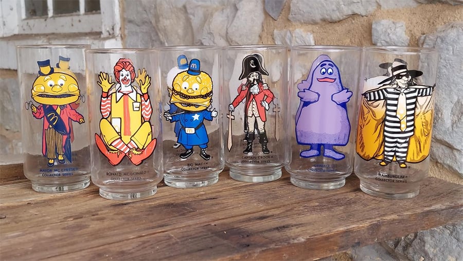 Six vintage McDonald's character glasses are lined up on a wooden shelf. The glasses feature Mayor McCheese, Ronald McDonald, Officer Big Mac, Captain Crook, Grimace, and the Hamburglar. Stone wall in the background.